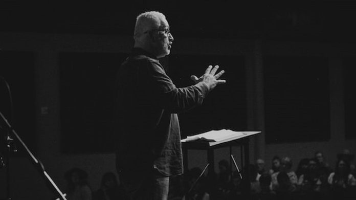 black and white phot of man with beard speaking to a crowd from a stage