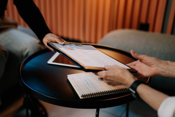 person handing cv, attached to a clipboard, accross desk